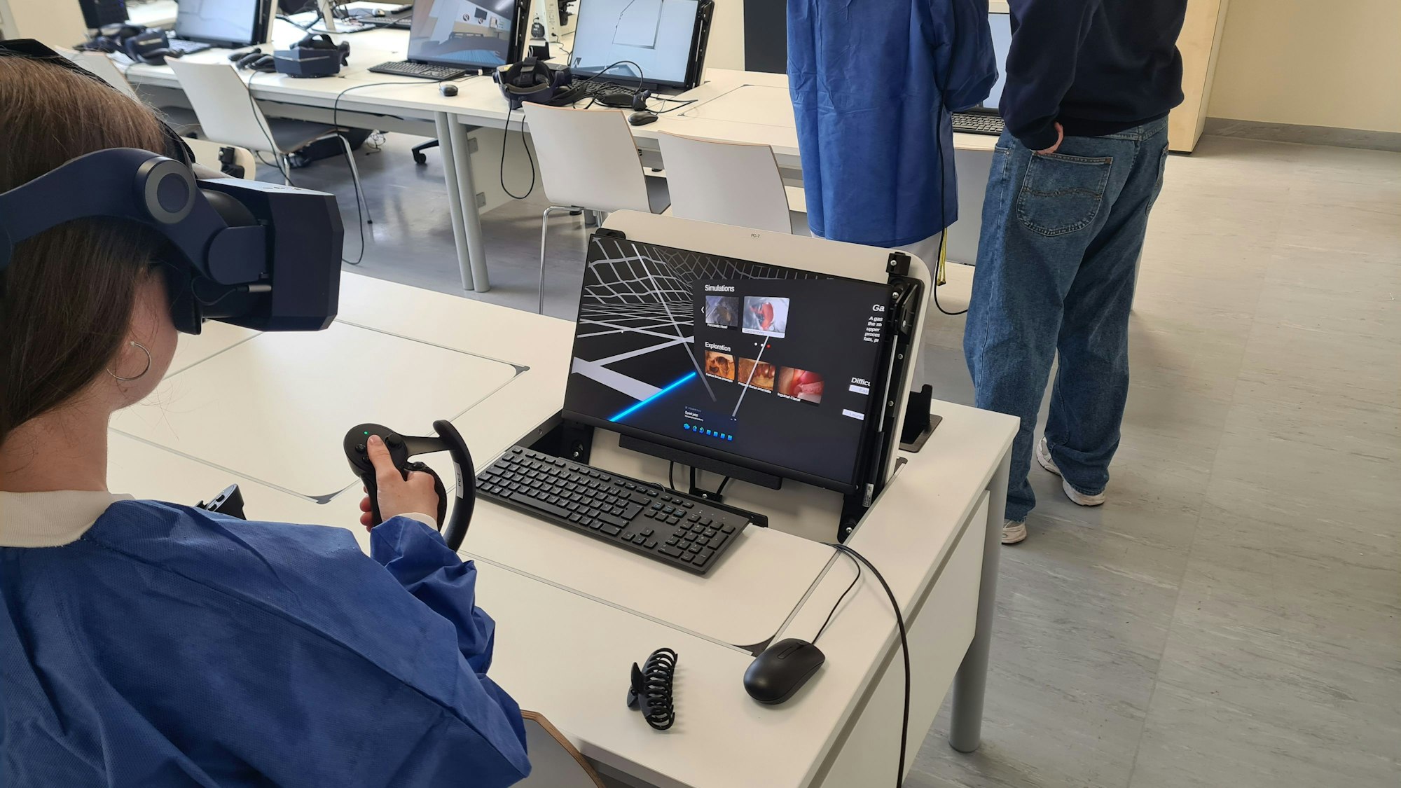 Eine Studentin an der Universität Köln sitzt an einem Computer und trainiert mit einer VR-Brille sowie zwei Controllern in ihrer Hand einen operativen Eingriff.