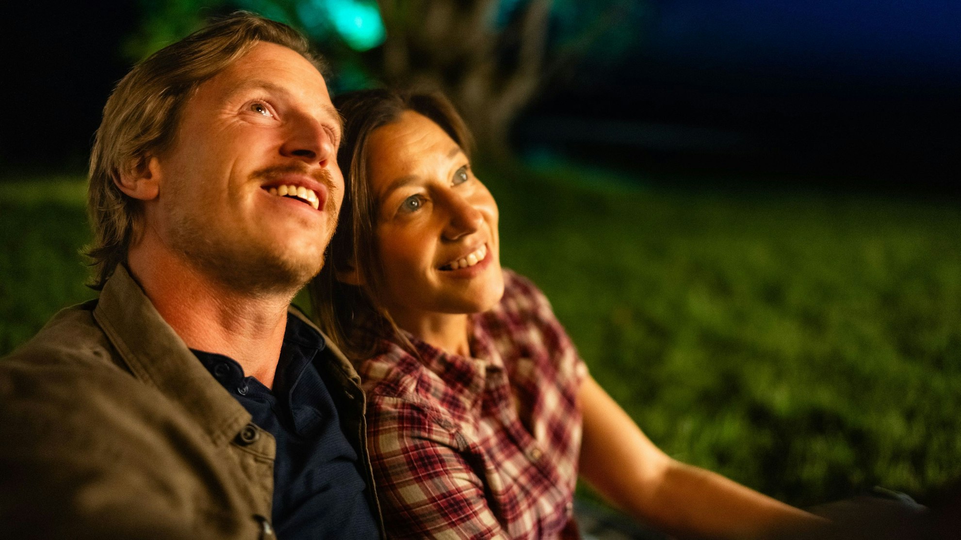 Cris Blohm und Hanno Senoner sitzen nebeneinander auf einer Wiese und schauen dem Feuerwerk zu.