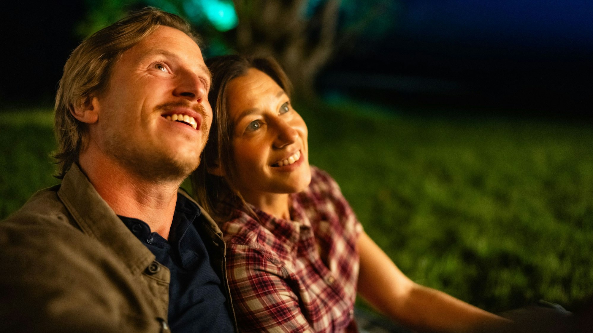 Cris Blohm und Hanno Senoner sitzen nebeneinander auf einer Wiese und schauen dem Feuerwerk zu.