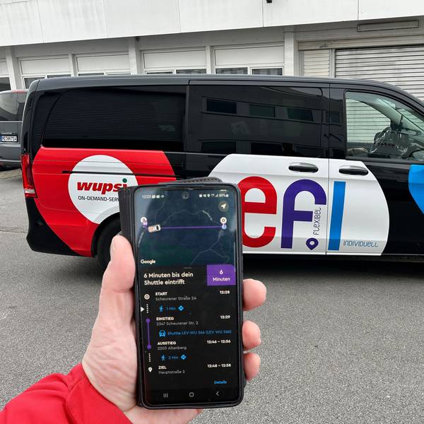 Eine Hand mit einem Smartphone und der „Efi“-App ist vor einem Auf-Abruf-Fahrzeug der „Efi“-Flotte zu sehen.