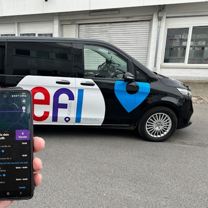 Eine Hand mit einem Smartphone und der „Efi“-App ist vor einem Auf-Abruf-Fahrzeug der „Efi“-Flotte zu sehen.