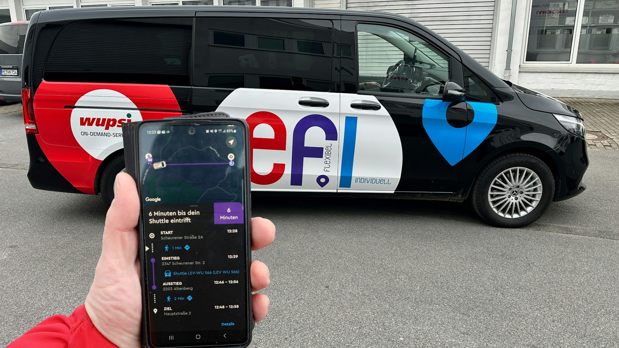 Ein „Efi“-Fahrzeug steht auf einem Parkplatz. Eine Hand hält davor ein Smartphone mit der „Efi“-Bestell-App.