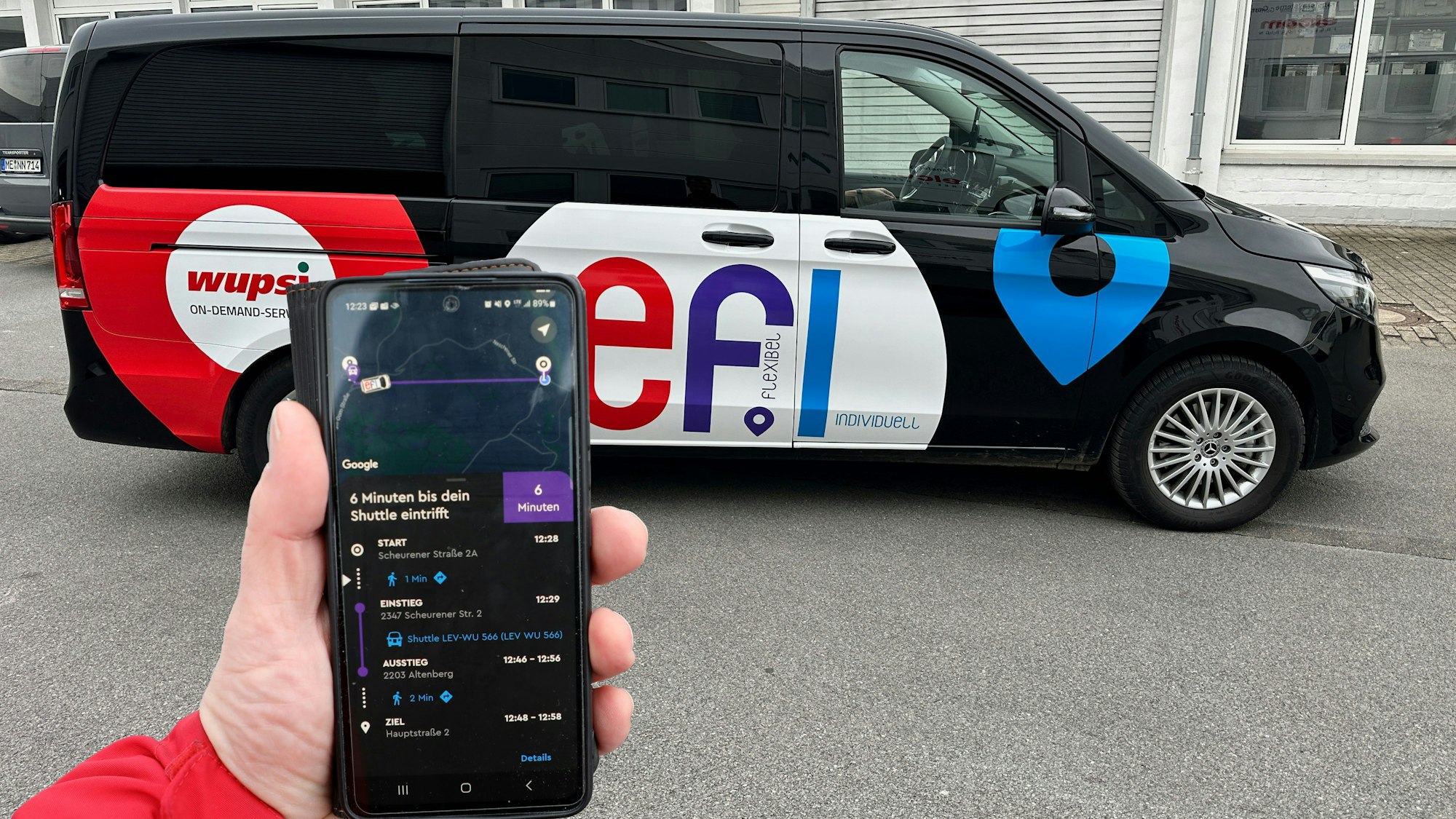 Eine Hand mit einem Smartphone und der „Efi“-App ist vor einem Auf-Abruf-Fahrzeug der „Efi“-Flotte zu sehen.