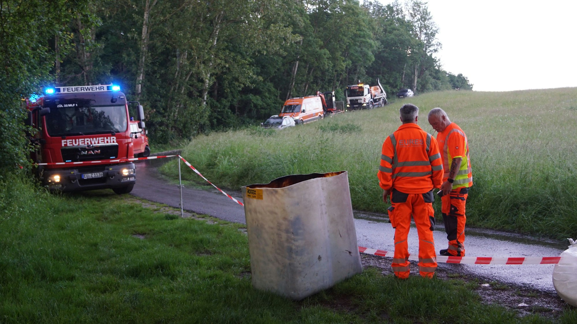 Auf dem Kunkel-Pütz-Weg sind die Einsatzfahrzeuge der Feuerwehr geparkt. Im Vordergrund warten zwei Mitarbeiter einer alarmierten Entsorgungsfirma.