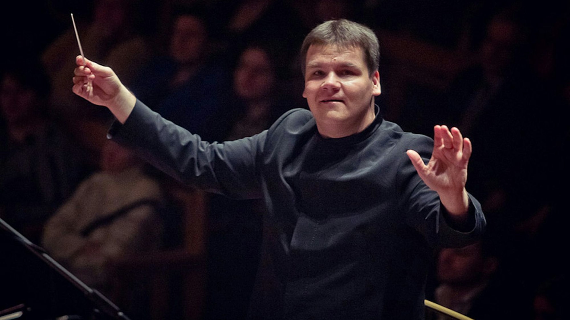 Andris Poga steht am Dirigentenpult, in der rechten Hand hält er den Taktstock, die linke Hand hat er angehoben.