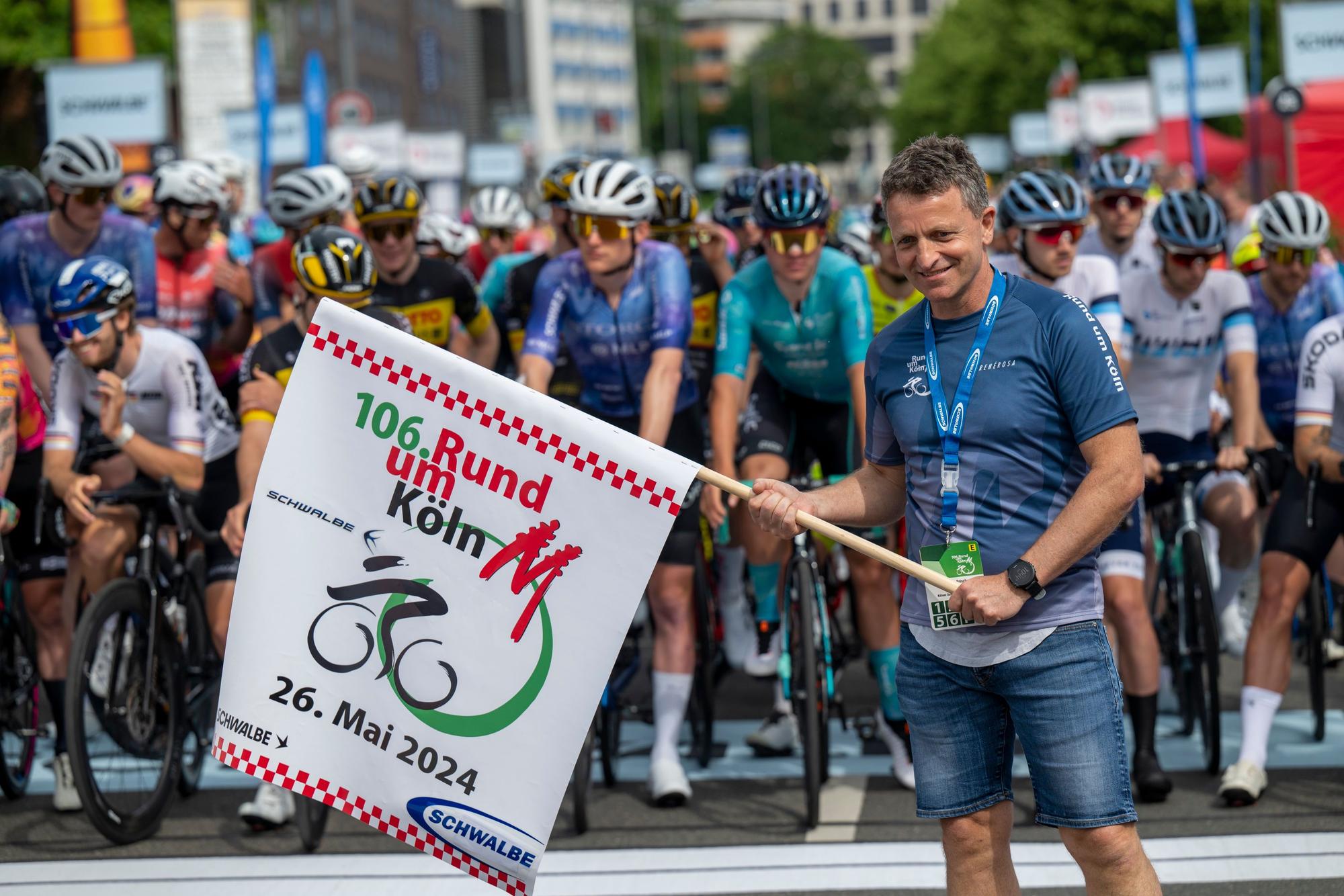 Ein Mann hält eine Flagge, hinter ihm warten Rennradfahrer.