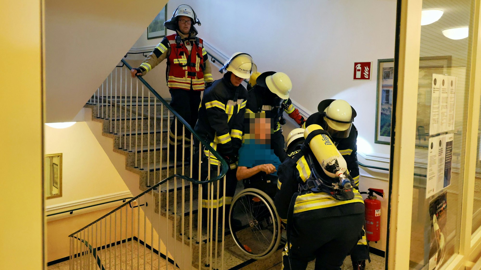 Vier Feuerwehrleute bugsieren den Mann in seinem Rollstuhl die Treppe hinab, ein Gruppenführer dahinter schaut zu.