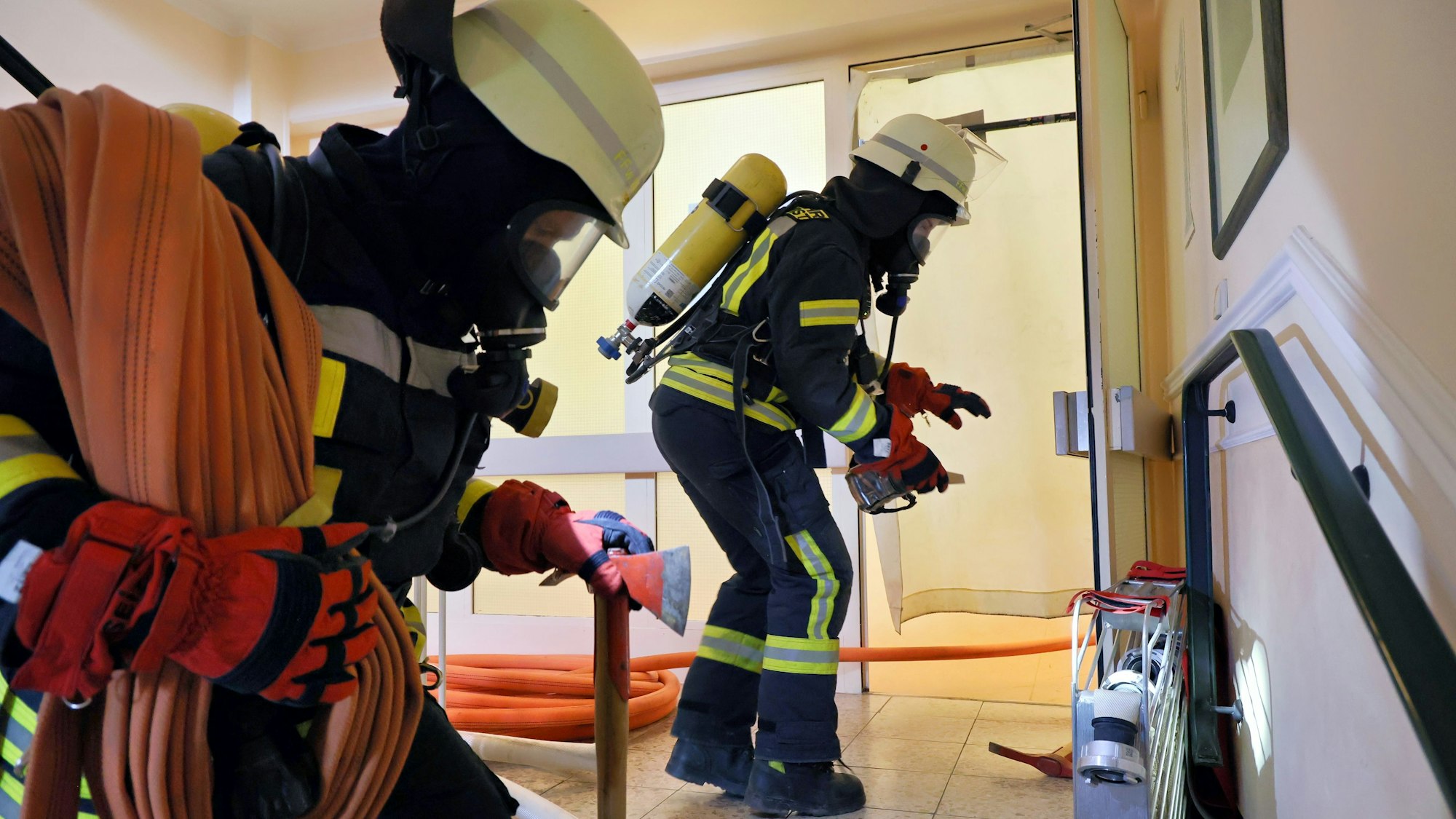 Die beiden Feuerwehrleute, kurz bevor sie unter dem Rauchverschluss durchschlüpfen. Beide tragen Atemschutz. Der hintere hat eine Feuerwehraxt in einer Hand und Schlauchmaterial geschultert.