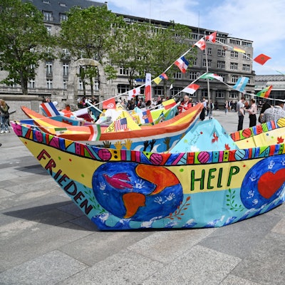 Zu sehen ist ein großes, bemaltes Papierfaltboot, das auf der Domplatte in Köln steht.