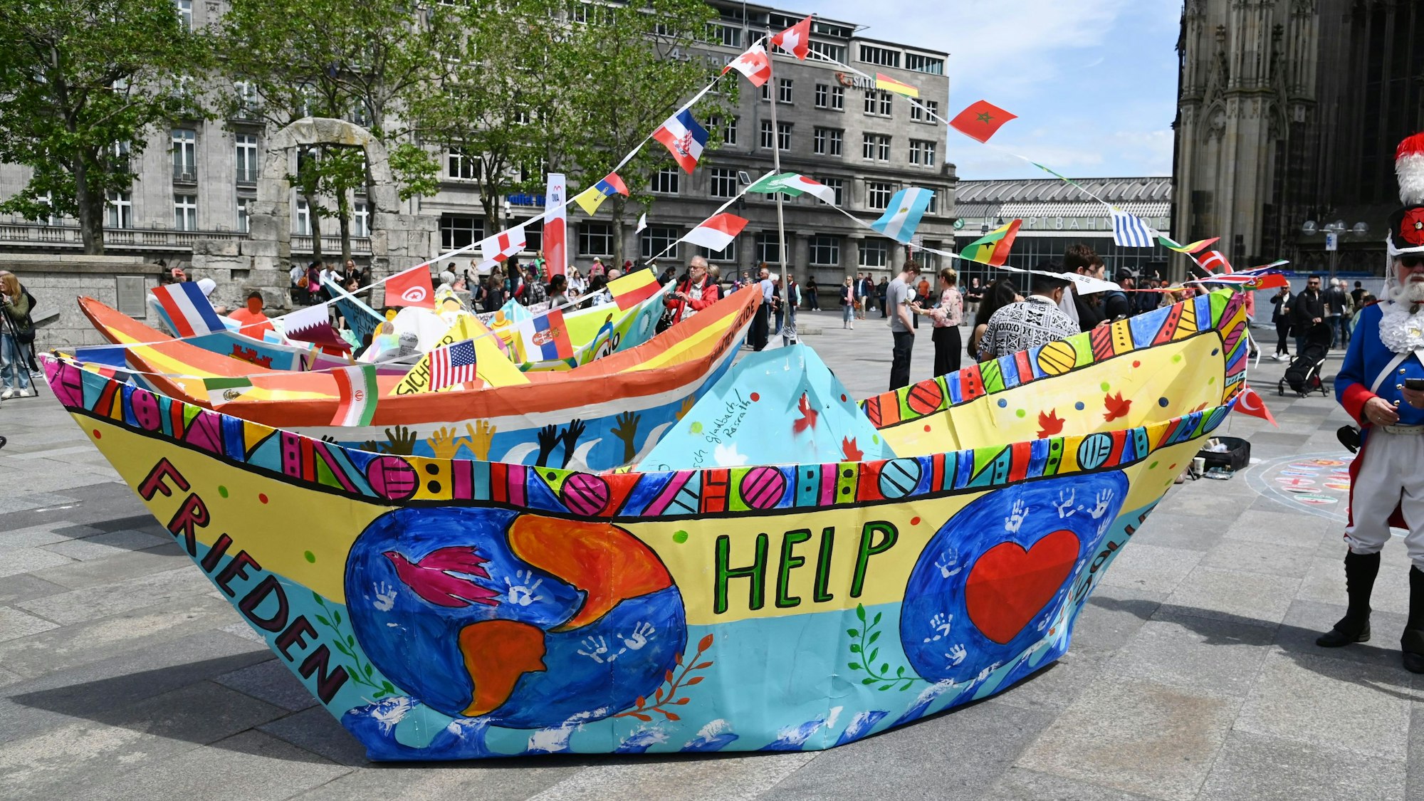 Zu sehen ist ein großes, bemaltes Papierfaltboot, das auf der Domplatte in Köln steht.