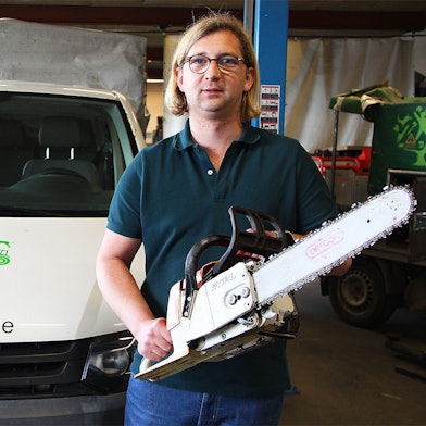 Sven Bung steht mit einer Motorsäge in einer Firmenhalle, links neben ihm ein Firmenwagen mit der Aufschrift „www.vitaforst.de“