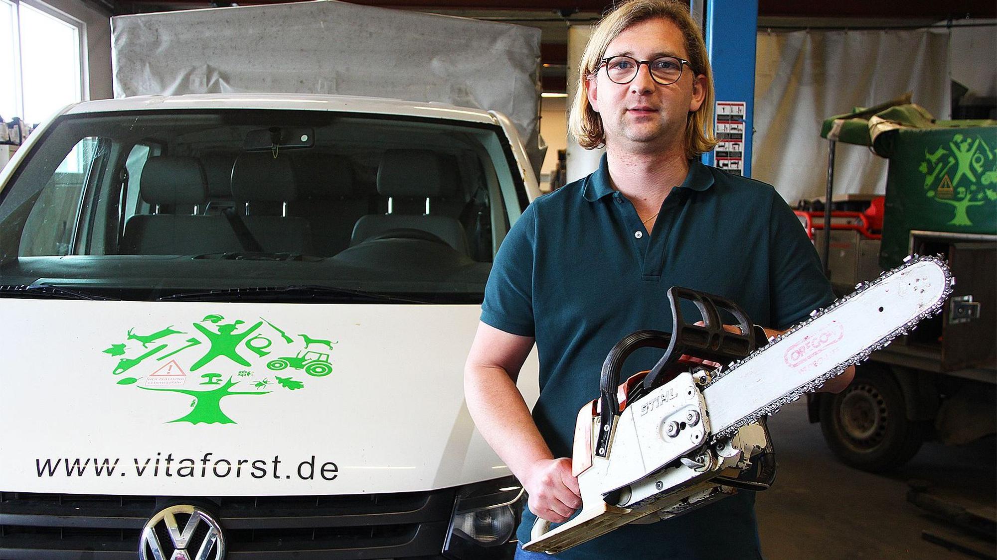 Sven Bung steht mit einer Motorsäge in einer Firmenhalle, links neben ihm ein Firmenwagen mit der Aufschrift „www.vitaforst.de“