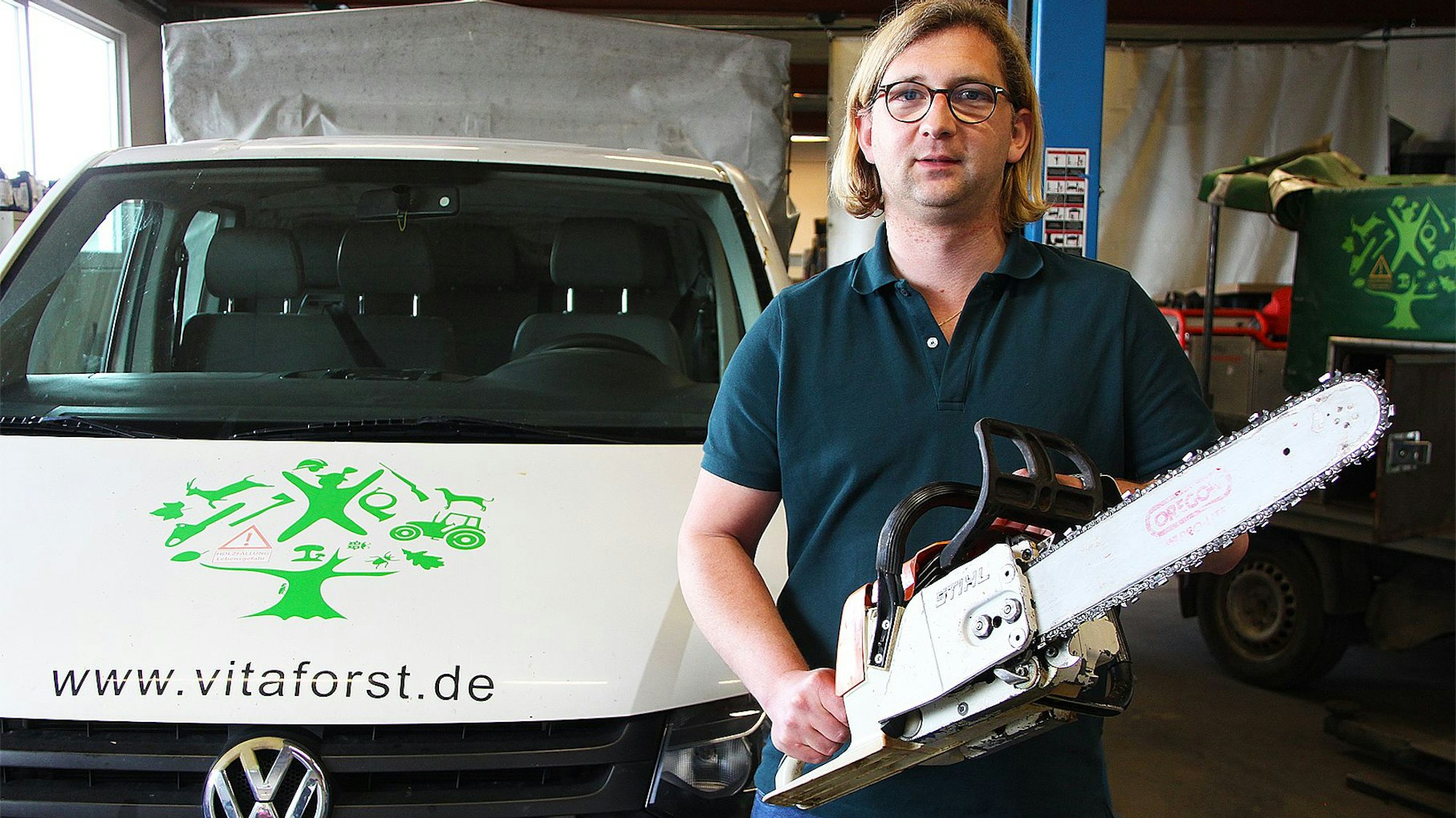 Sven Bung steht mit einer Motorsäge in einer Firmenhalle, links neben ihm ein Firmenwagen mit der Aufschrift „www.vitaforst.de“