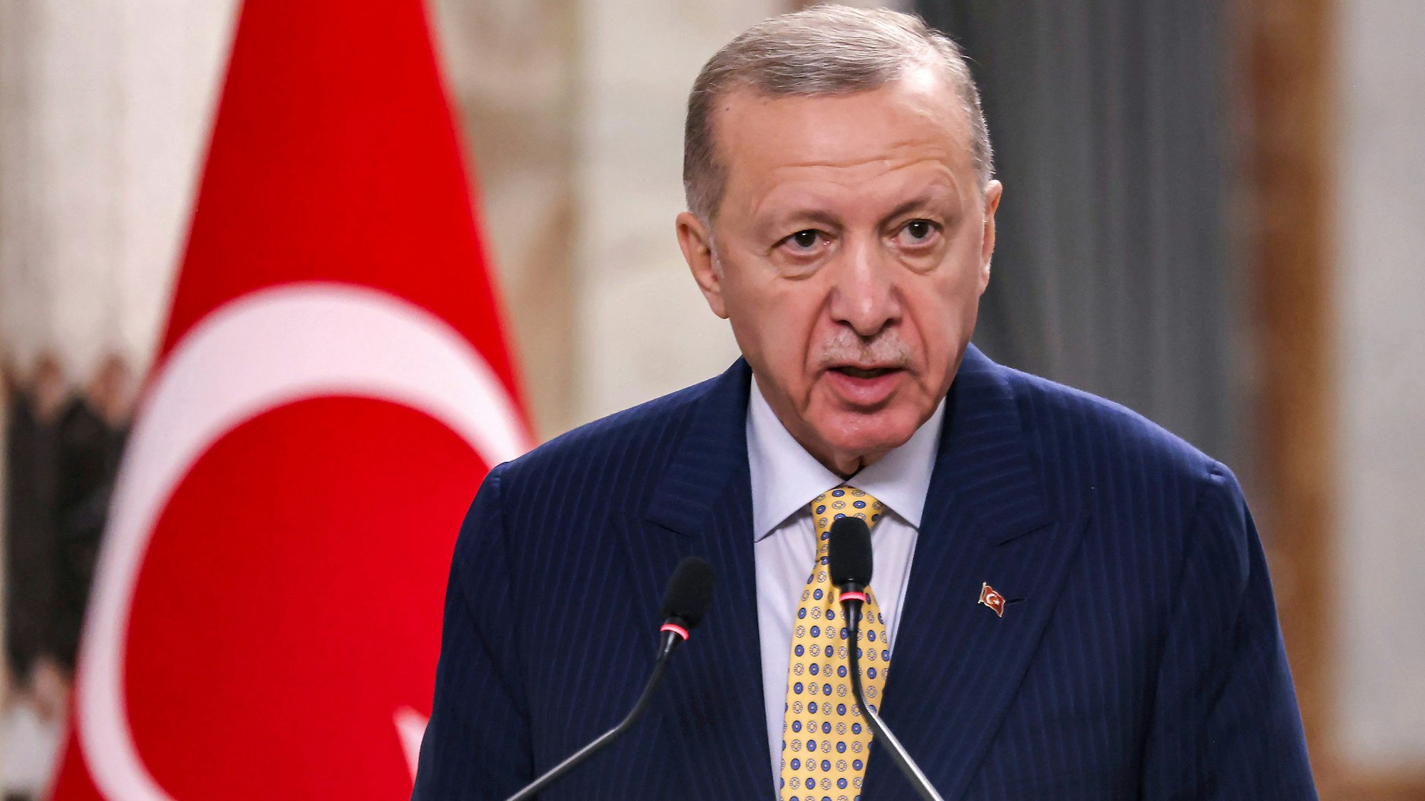 Der türkische Präsident Recep Tayyip Erdogan spricht in zwei Mikrofone. Im Hintergrund ist eine Flagge der Türkei unscharf gestellt.