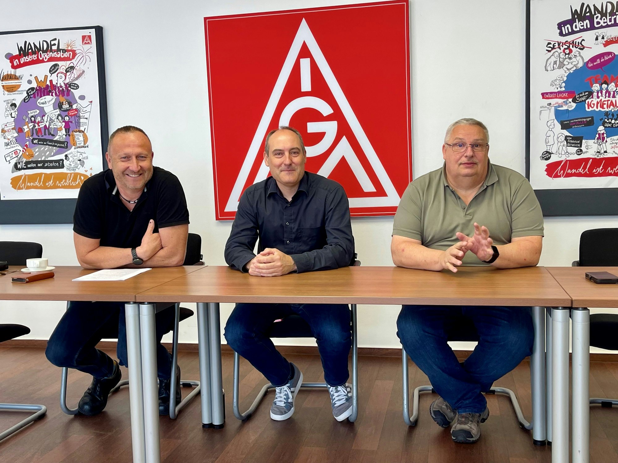 Drei Männer sitzen an einem Tisch, hinter ihnen hängt das rote IG-Metall-Logo.