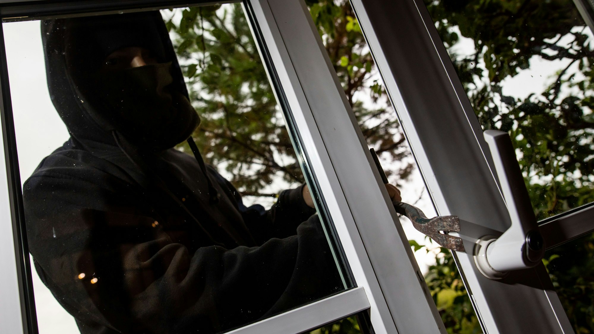 Ein Mann mimt einen Einbrecher und benutzt ein Brecheisen, um eine gekippte Terrassentür aufzuhebeln.