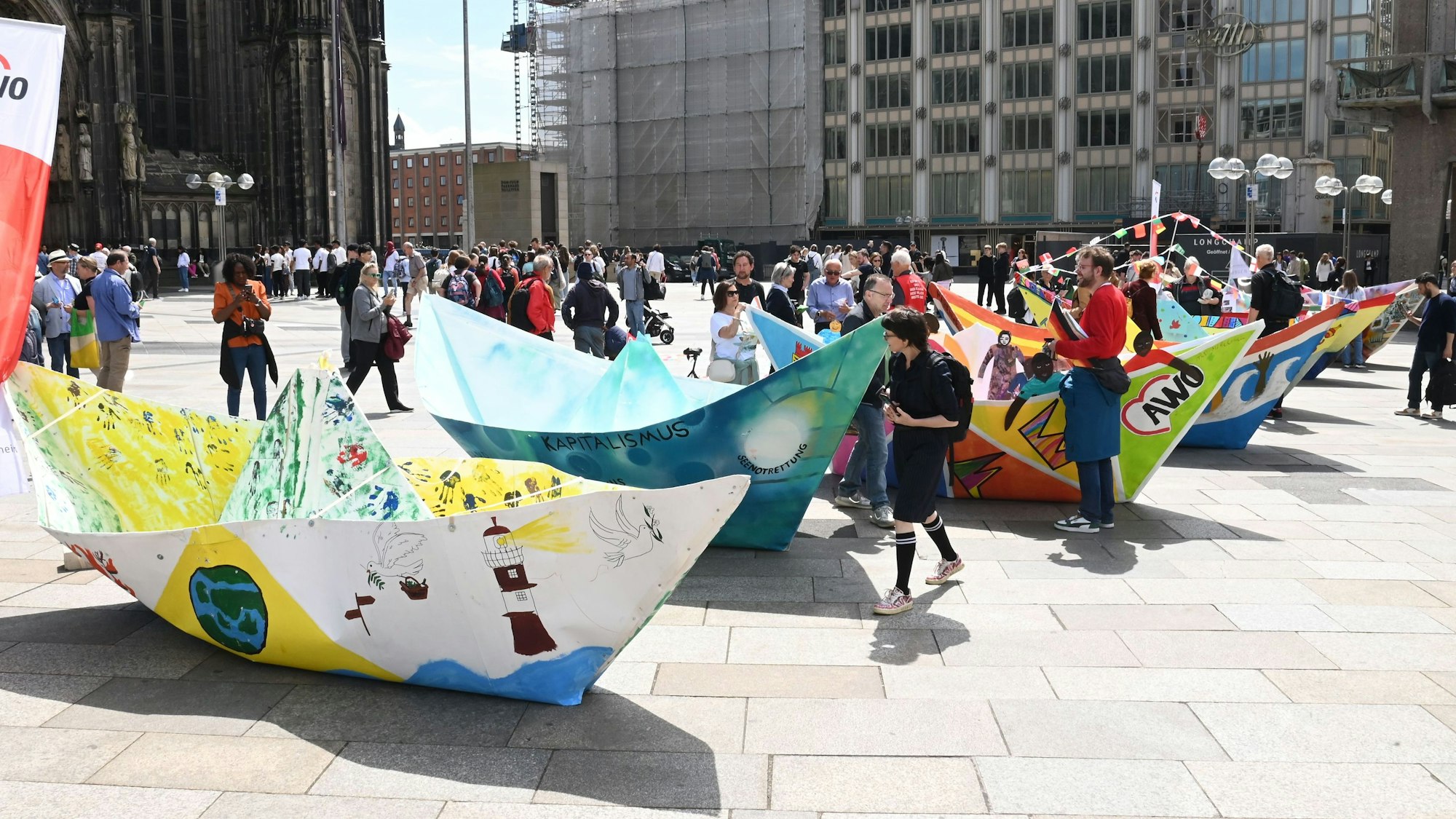 Zu sehen sind mehrere bemalte Papierfaltboote, die auf der Domplatte in Köln stehen.