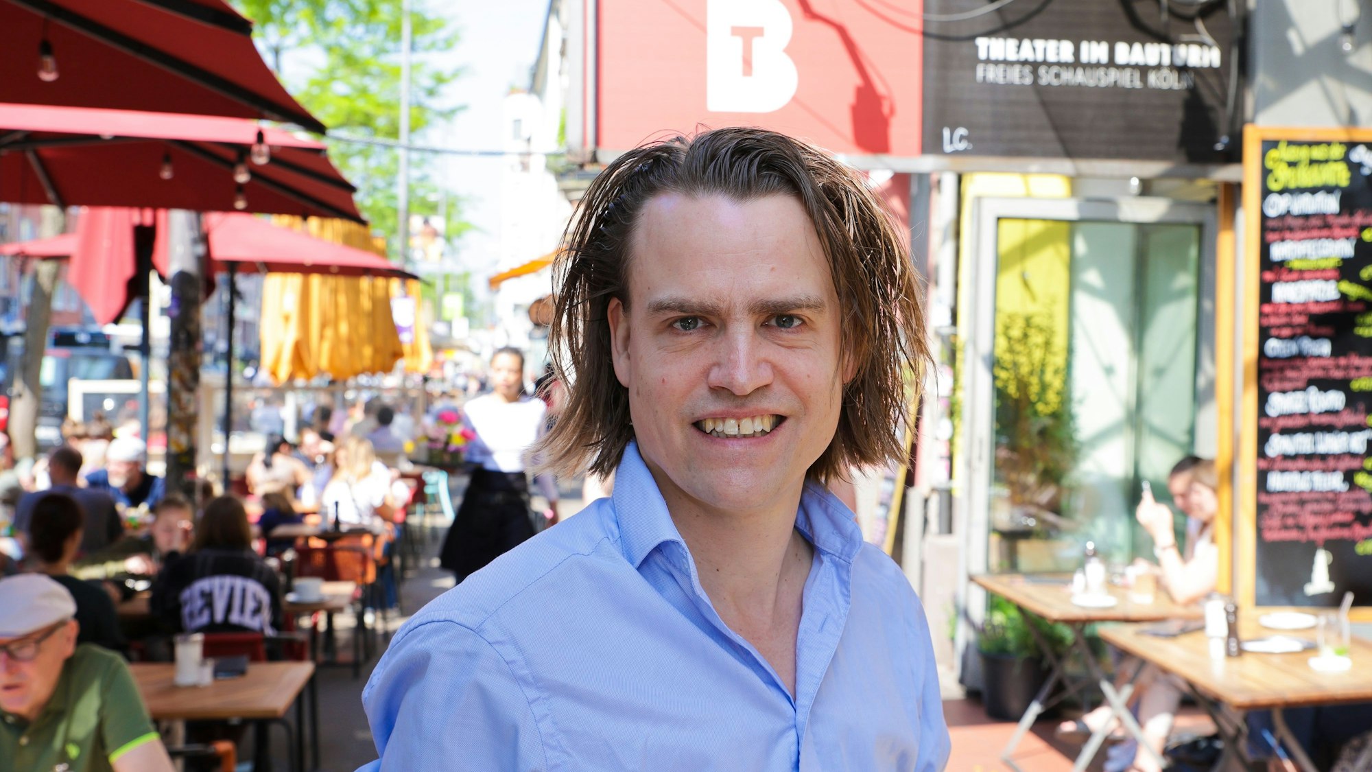 Das Bild zeigt Laurenz Leky an der Aachener Straße. Seine langen Haare gehen ihm bis zum Kinn. Er trägt ein hellblaues Hemd. Im Hintergrund ein Banner mit der Aufschrift: Theater im Bauturm, Freies Schauspiel Köln.