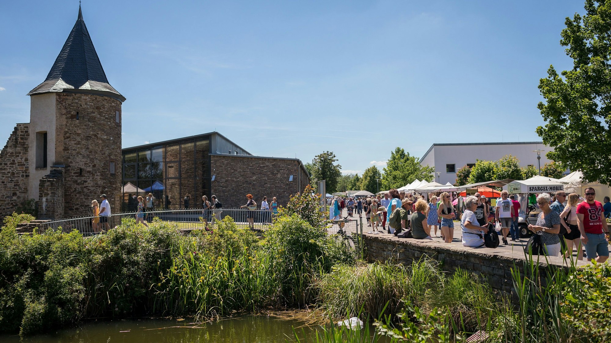 Ein Archivbild zeigt den Wollmarkt auf dem Museumsgelände – mit Publikum, Verkaufsständen, einem Teich im Vordergrund und dem Gästehaus Mottenburg im Hintergrund.