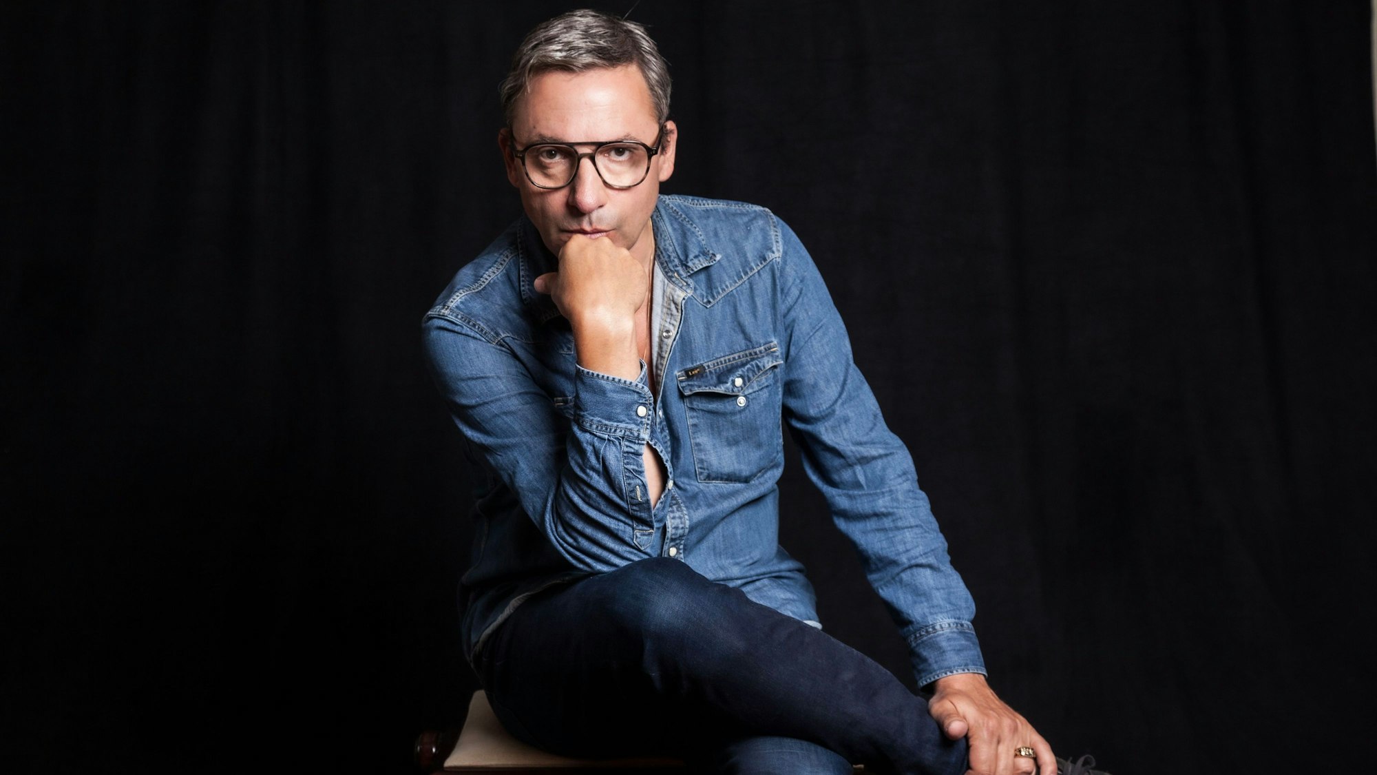 Zu sehen ist Rocko Schamoni, sitzend vor einem schwarzen Hintergrund.