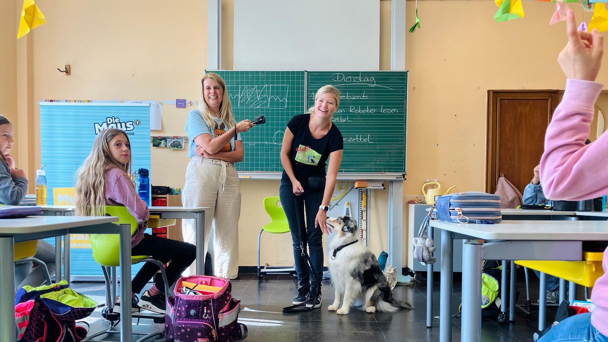 Maus-Reporterin Christina Pannhausen, Hundetrainerin Stephanie Maron und die Hündin Fly stehen vor der Klasse. Pannhausen hat ein Aufnahmegerät in der Hand.