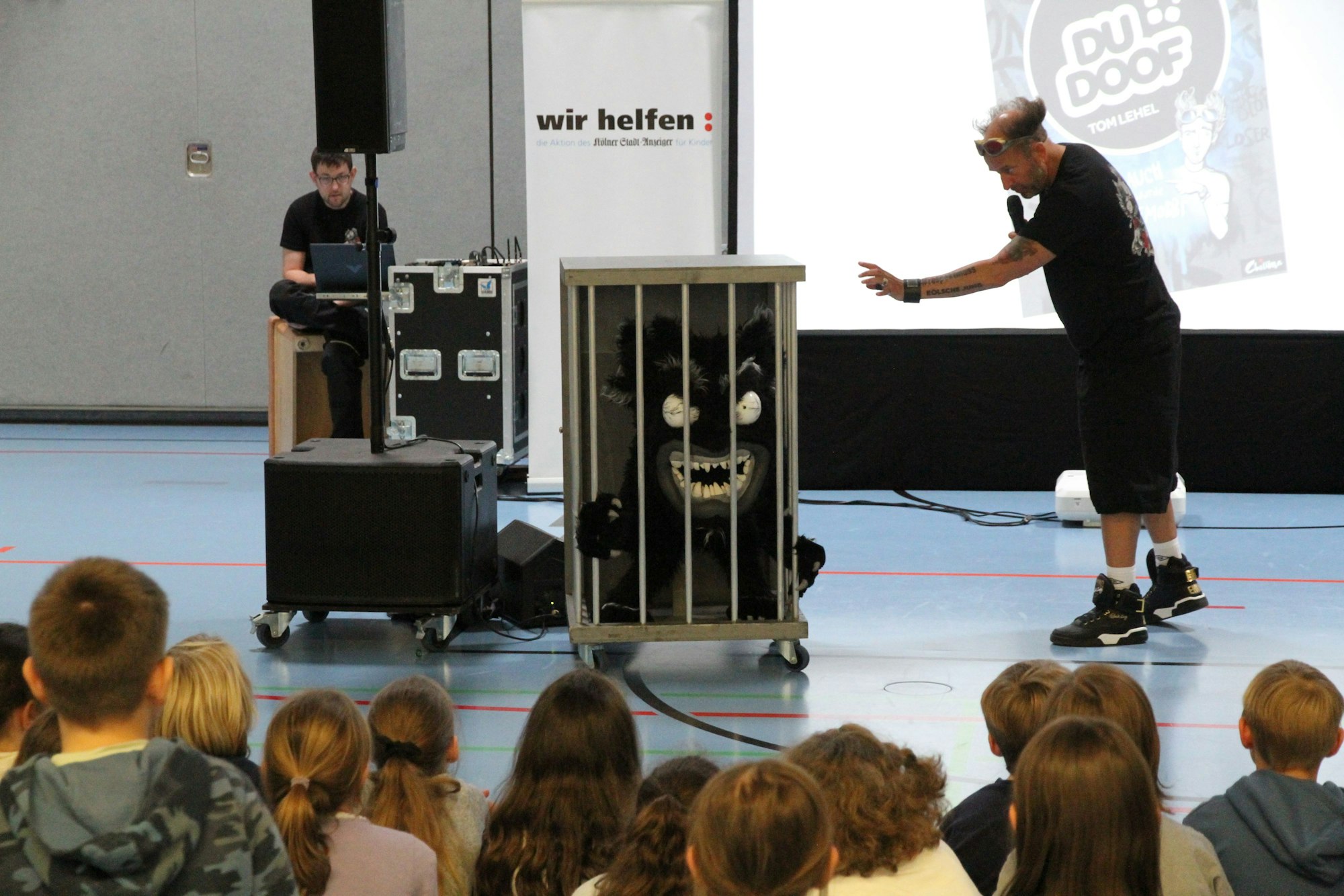 Ein Mann vor Schülern in einer Turnhalle mit einer Monsterpuppe in einem Käfig