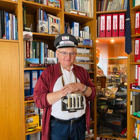 Bernhad Geuß steht vor einem Regal. Auf dem Kopf trägt er eine Schaffnermütze, um den Hals hängt ihm ein Galoppwechsler.