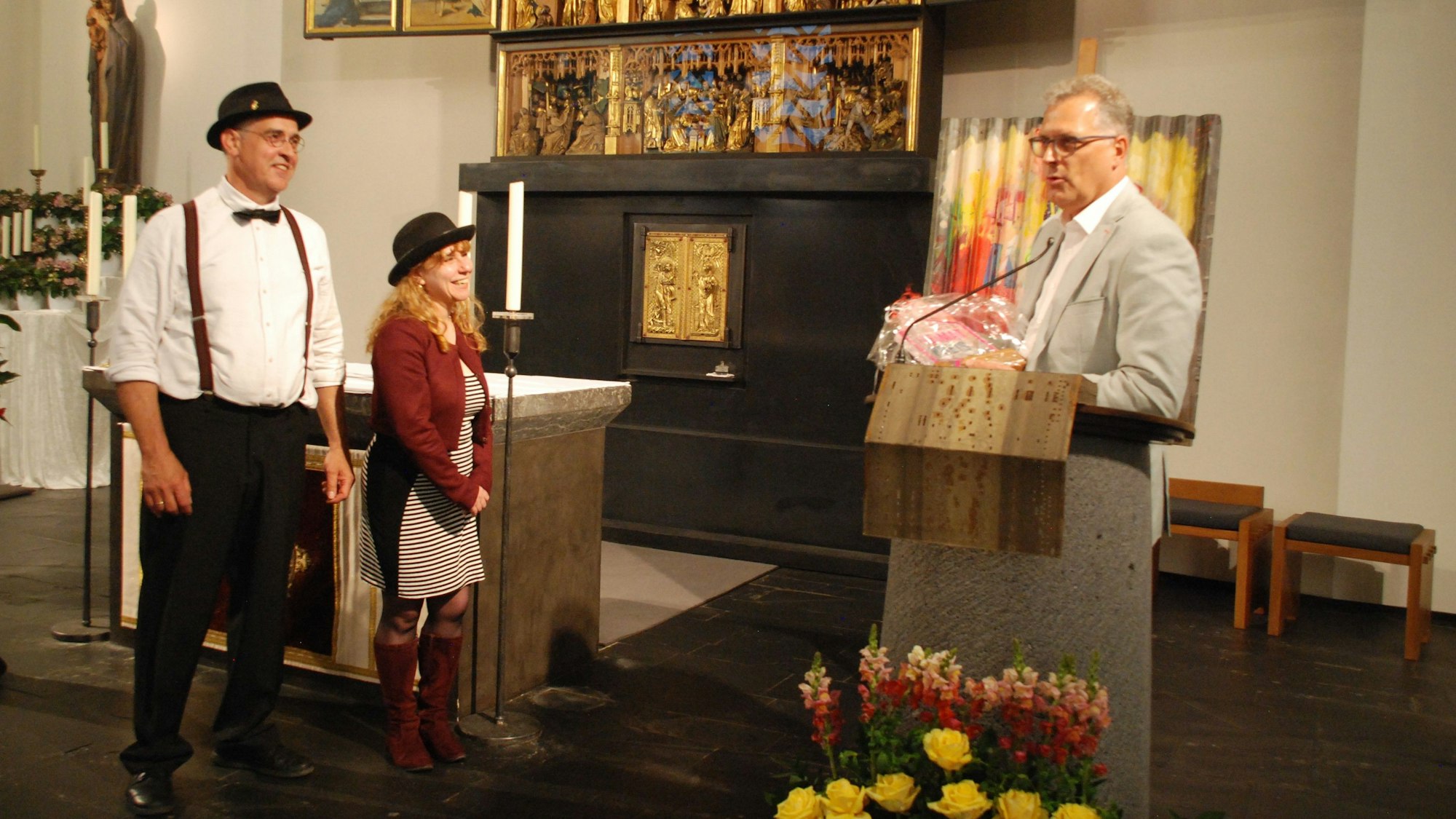 Am Rednerpult in einer Kirche steht Zülpichs Bürgermeister Ulf Hürtgen. Er hält ein Geschenk zum Heimatpreis in der Hand, den er Toni Schleifer und Eugenia Fabrizi verleihen wird. Die beiden stehen links neben ihm.