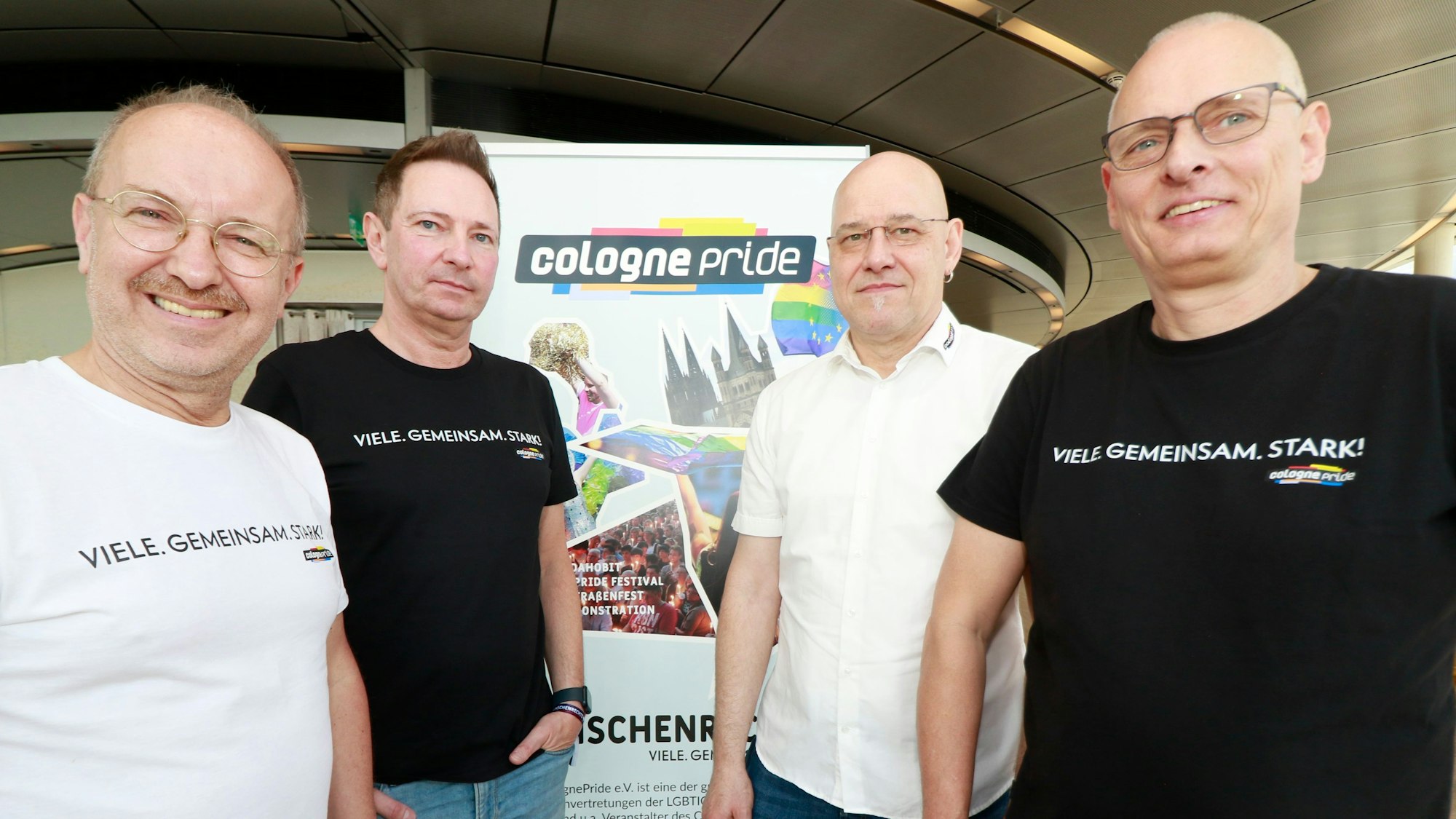 Zu sehen sind vier Männer, die vor einem Plakat mit der Aufschrift „cologne pride“ stehen.