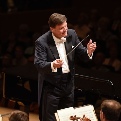 Das Bild zeigt Christian Thielemann, er trägt einen schwarzen Anzug mit weißem Hemd und hält einen Taktstock in der Hand. Er ist seinem Orchester zugewandt.
