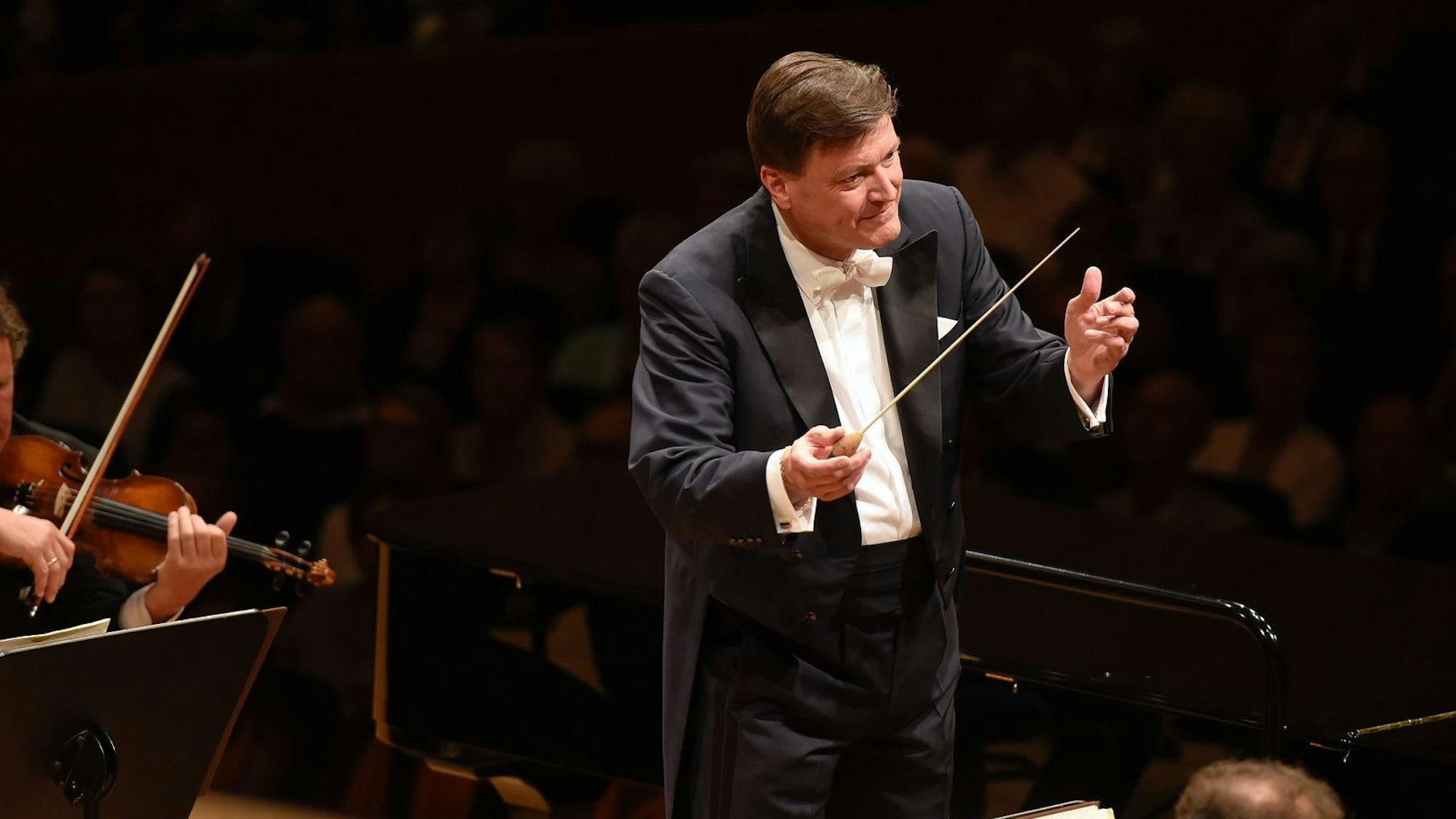 Das Bild zeigt Christian Thielemann, er trägt einen schwarzen Anzug mit weißem Hemd und hält einen Taktstock in der Hand. Er ist seinem Orchester zugewandt.