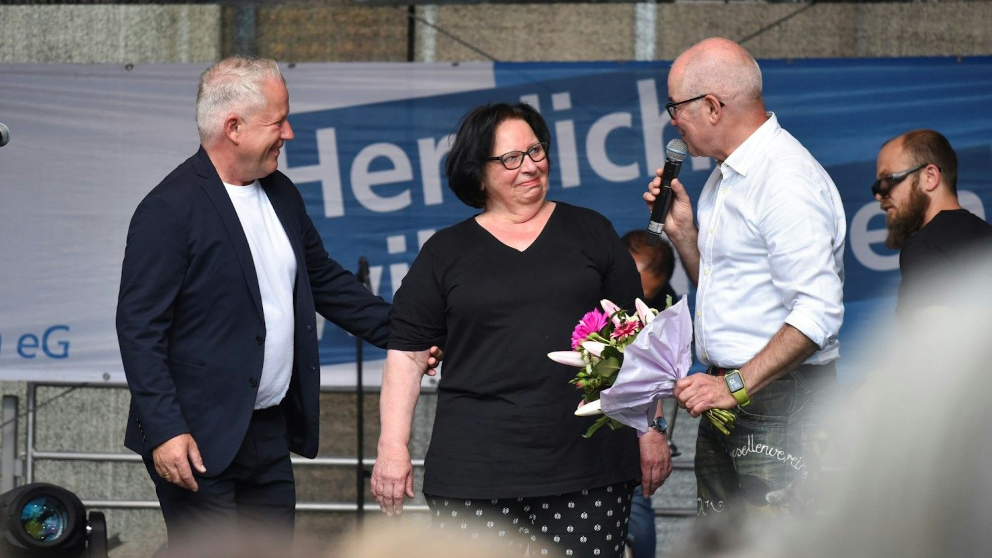 Eine Frau bekommt Blumen von einem Mann in weißem Hemd. Sie trägt ein schwarzes T-Shirt. Neben der Frau steht ein weiterer Mann, er trägt ein weißes T-Shirt und ein blaues Sakko.