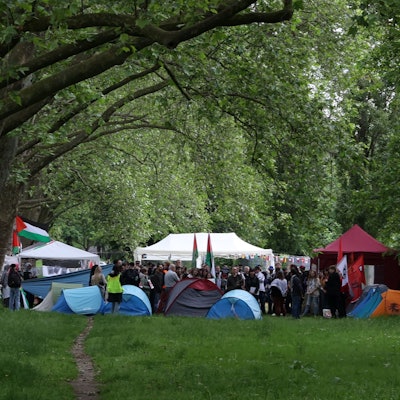 Zelte stehen zusammen auf einer Wiese, links ist eine Fahne von Palästina zu sehen.