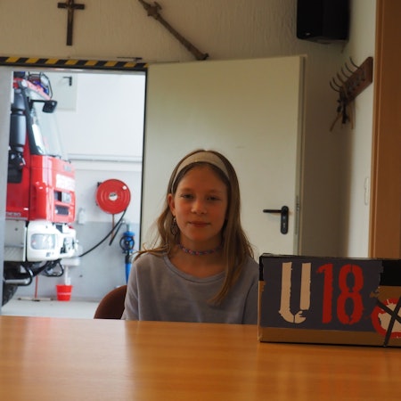Die Elfjährige sitzt vor ihrer selbstgebastelten Wahlurne mit der Aufschrift U18, im Hintergrund ein Löschfahrzeug der Feuerwehr.