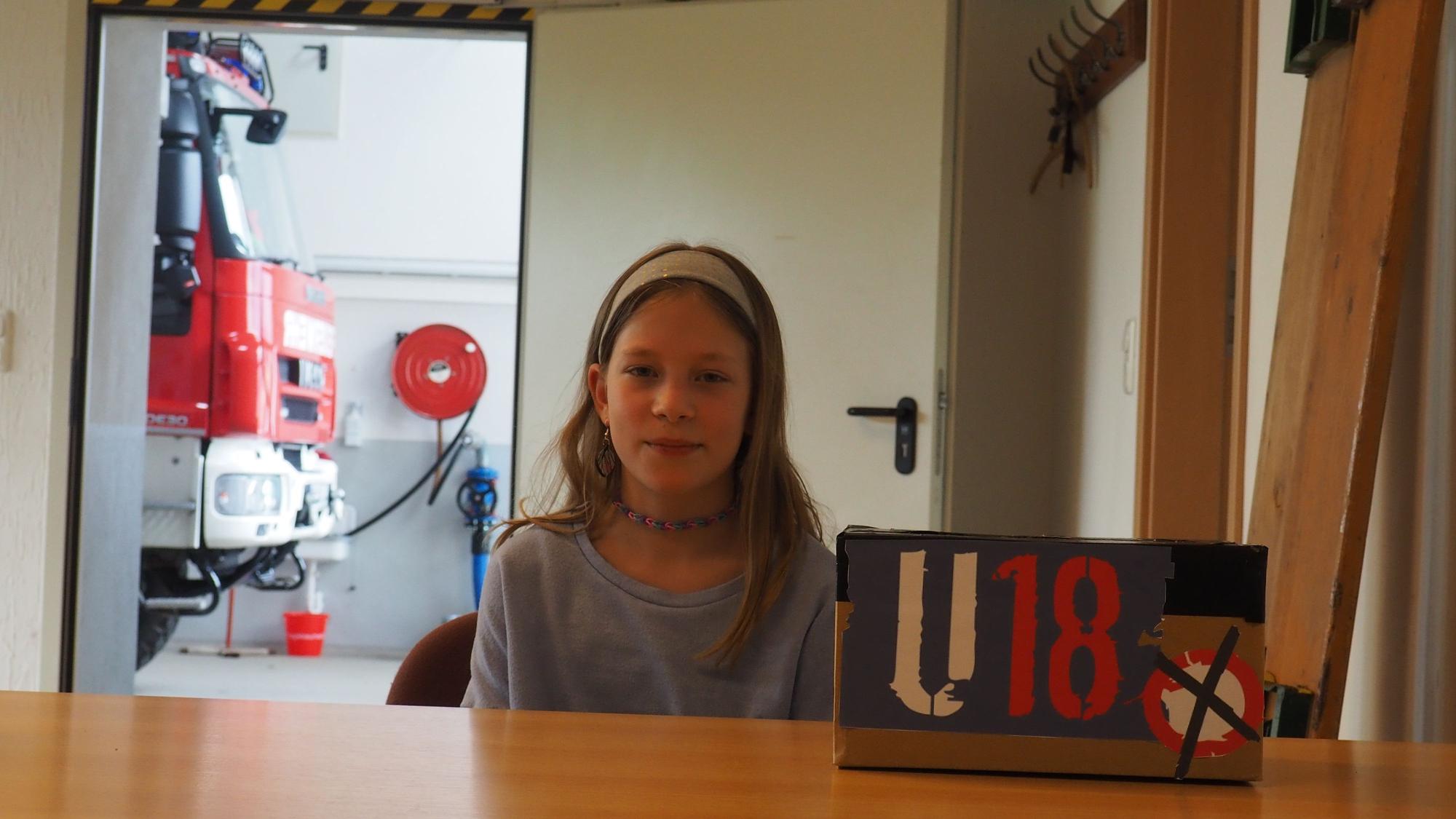 Die Elfjährige sitzt vor ihrer selbstgebastelten Wahlurne mit der Aufschrift U18, im Hintergrund ein Löschfahrzeug der Feuerwehr.