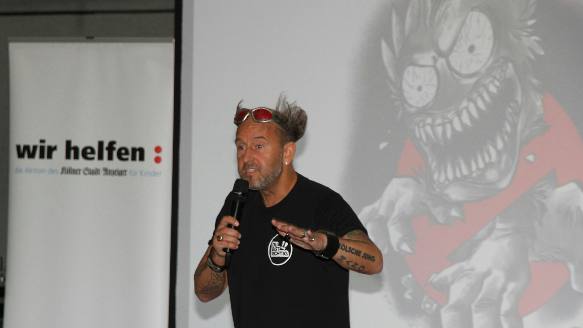 Ein Mann mit hoch gegelten Haaren und Mikrophon vor einer an die Wand projizierten Monsterfratze