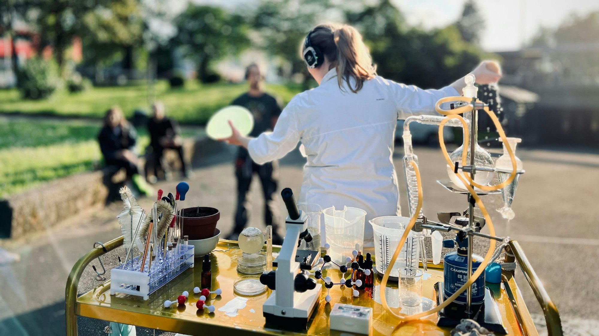 Auf dem Bild ist das mobile Labor zu sehen, ein kleiner Rollwagen mit allerlei wissenschaftlichen Instrumenten. Dahinter eine Schauspielerin, die einen weißen Kittel trägt und Kopfhörern aufhat.