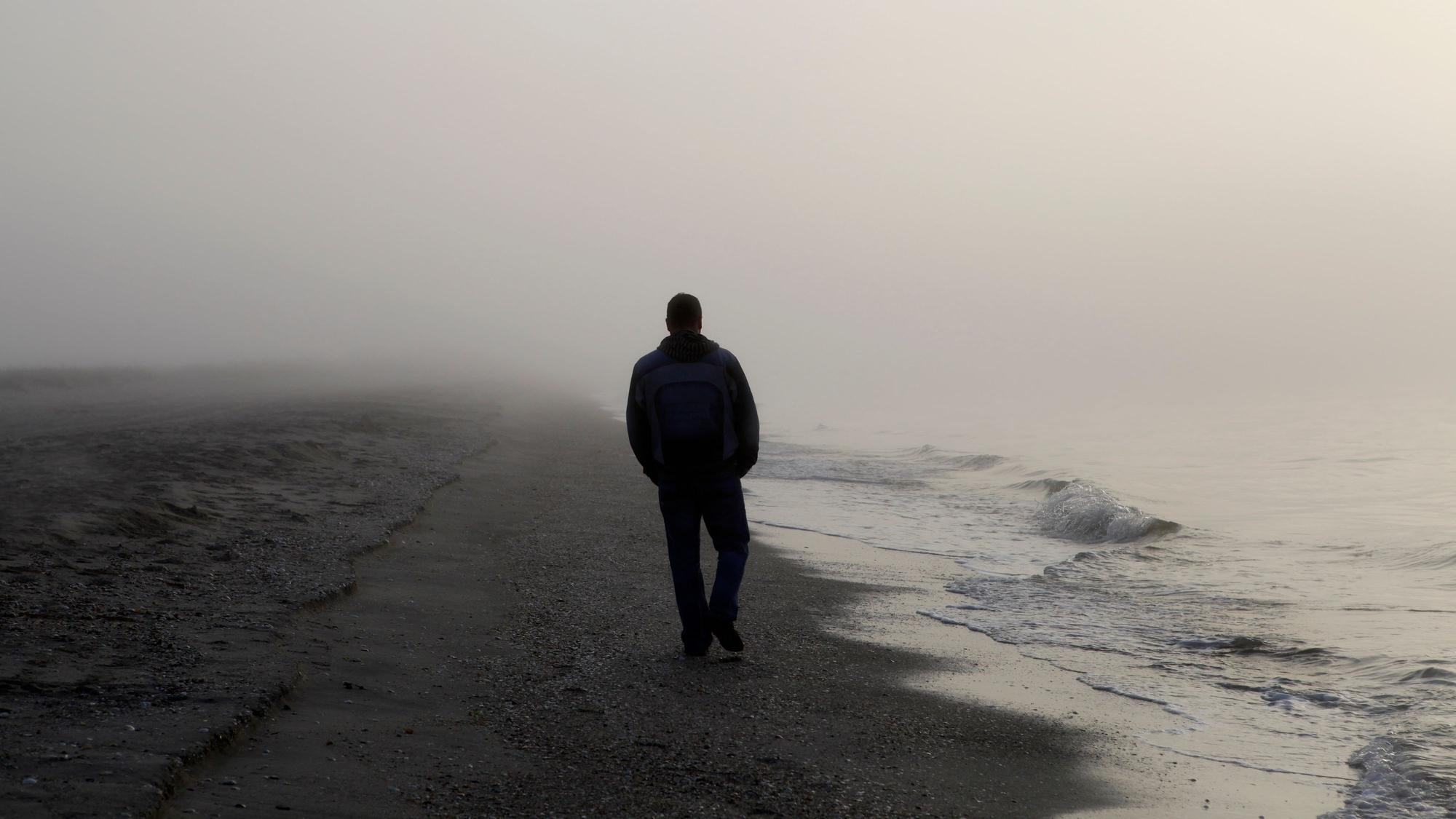 Ein Mann, von dem nur die Umrisse zu erkennen sind, spaziert an einem nebeligen Strand entlang. (Symbolbild)