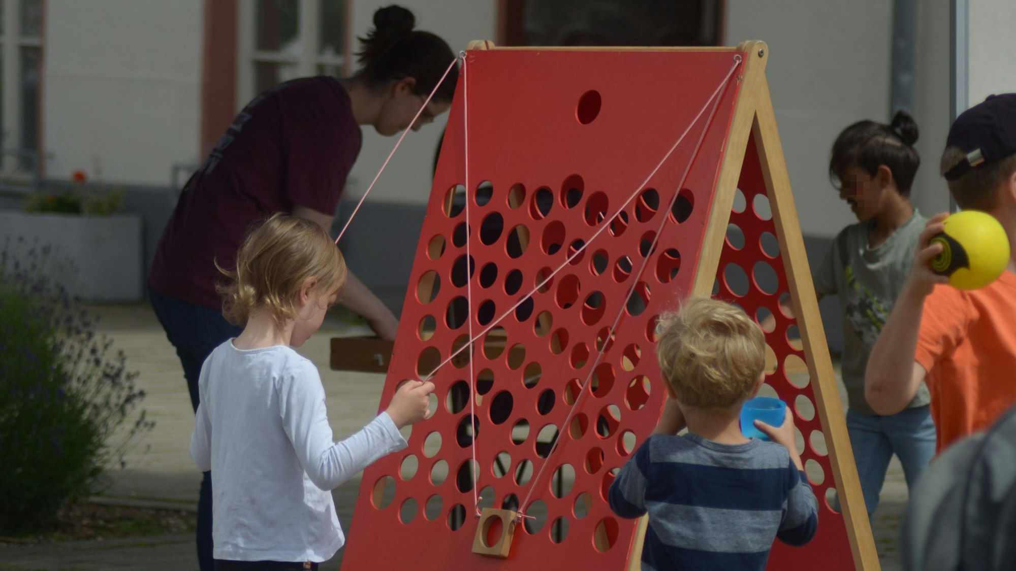 Ein Kind spielt mit einem Seilzugsystem an einer roten gelöcherten Tafel.