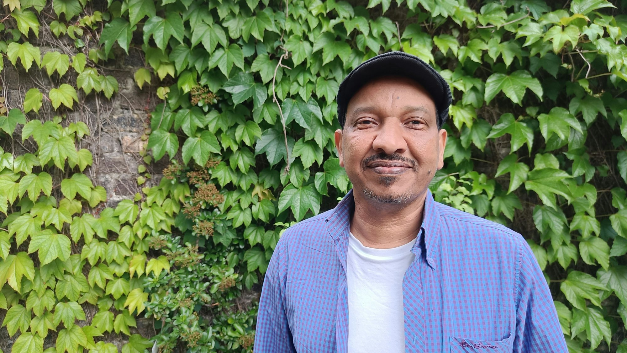 Ein Mann im blauen Hemd steht vor einer mit Grünpflanzen bewachsenen Wand.