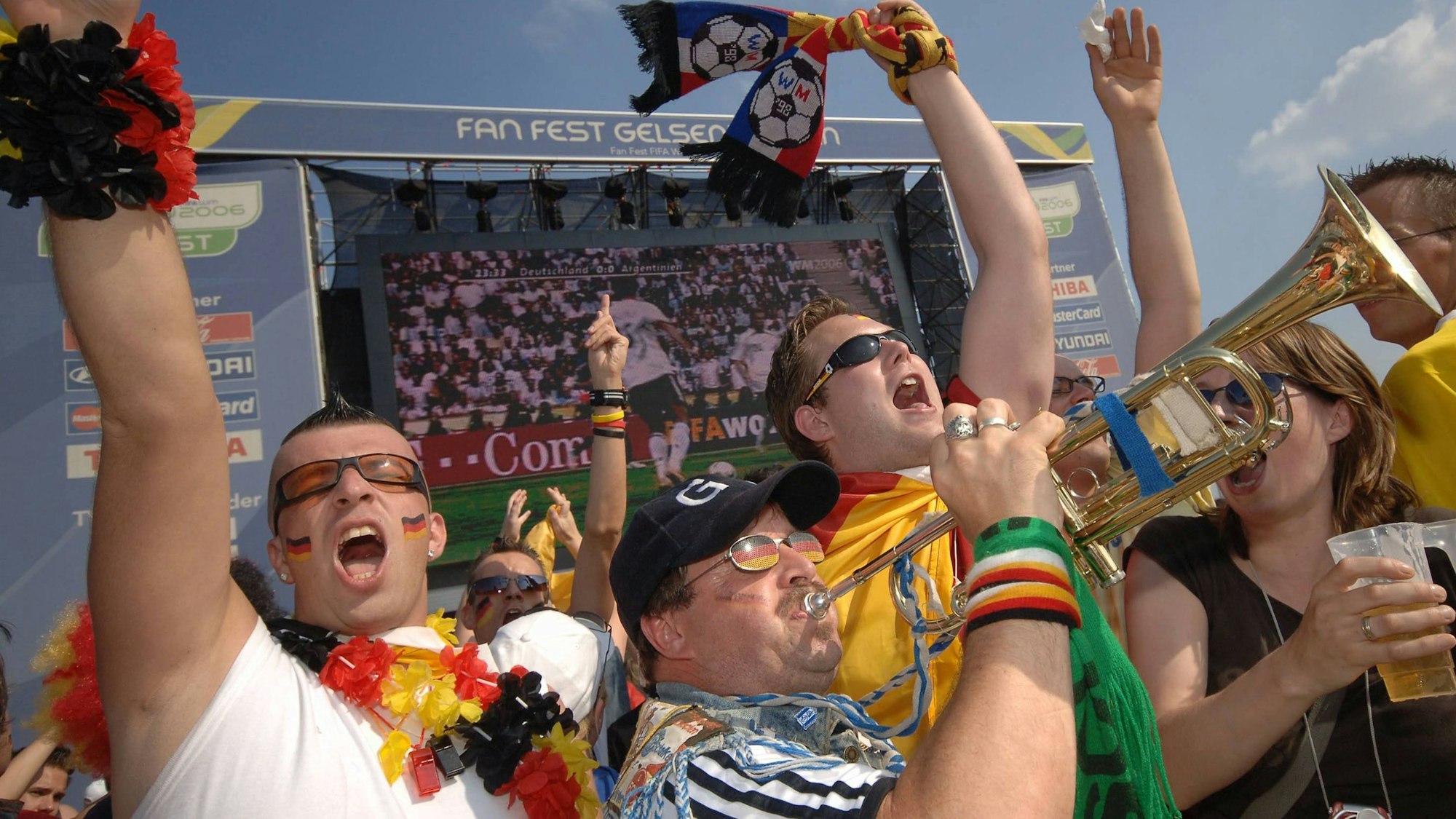 Das Bild zeigt feiernde Fans bei der WM 2006. Die Fans recken die Arme in die Lüfte, schwenken ihre Schals. Einer spielt Trompete. Im Hintergrund eine große Leinwand, die das Fußballspiel überträgt.