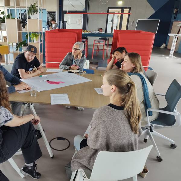 Das Foto zeigt eine der sieben Arbeitsgruppen, deren Mitglieder an einem Tisch Platz genommen haben. In der Mitte liegt ein großes Blatt, auf das die Gedanken und Ergebnisse geschrieben wurden.