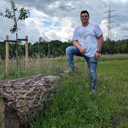 Ein Mann steht auf einer Wiese mit frisch gepflanzten Bäumen, ein Bein auf einen alten Baumstamm gestellt.