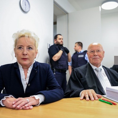 Zu sehen ist eine blonde Frau, die neben einem Mann mit Krawatte sitzt. Im Hintergrund stehen zwei Männer in einem Gerichtssaal.
