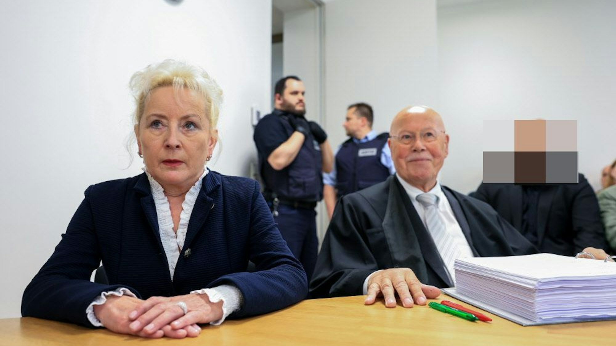 Zu sehen ist eine blonde Frau, die neben einem Mann mit Krawatte sitzt. Im Hintergrund stehen zwei Männer in einem Gerichtssaal.