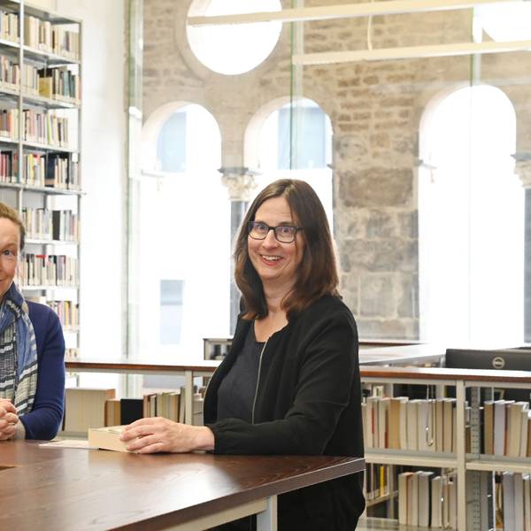 Die beiden Professorinnen sind in einer Bibliothek der Kunsthochschule für Medien Köln. Sie lehnen an einem hölzernen Tresen, hinter ihnen sind Regale mit Büchern.