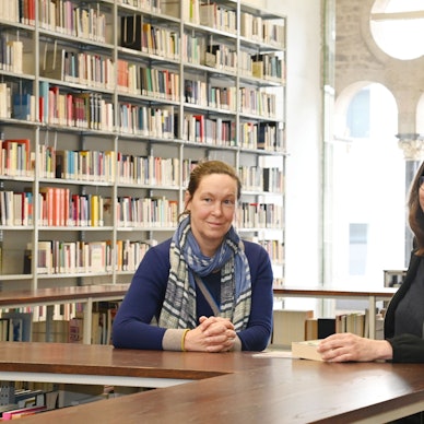 Die beiden Professorinnen sind in einer Bibliothek der Kunsthochschule für Medien Köln. Sie lehnen an einem hölzernen Tresen, hinter ihnen sind Regale mit Büchern.