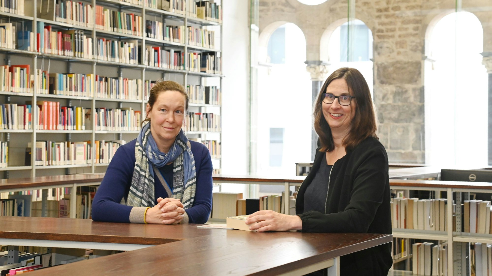 Die beiden Professorinnen sind in einer Bibliothek der Kunsthochschule für Medien Köln. Sie lehnen an einem hölzernen Tresen, hinter ihnen sind Regale mit Büchern.