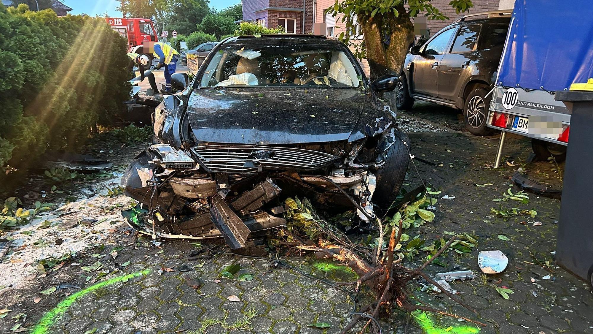Ein demoliertes Auto steht nach einem Unfall auf einem Hof, davor liegen von einem Baum abgerissene Äste. Im Hintergrund ist ein Feuerwehrfahrzeug zu sehen.