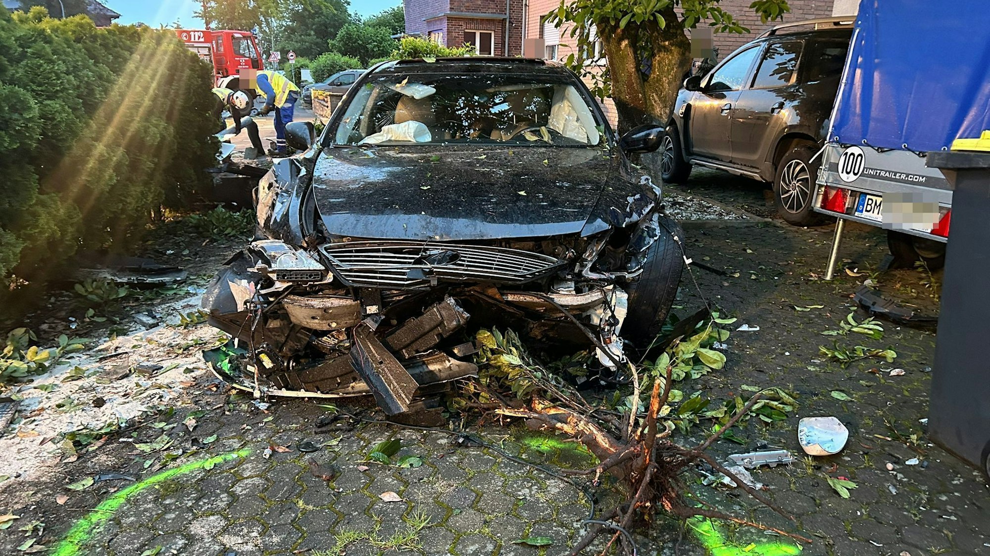 Ein demoliertes Auto steht nach einem Unfall auf einem Hof, davor liegen von einem Baum abgerissene Äste. Im Hintergrund ist ein Feuerwehrfahrzeug zu sehen.